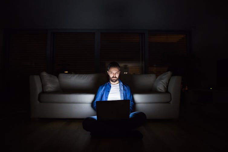 A man in a dark room is sitting on the floor using a laptop while leaning on a sofa