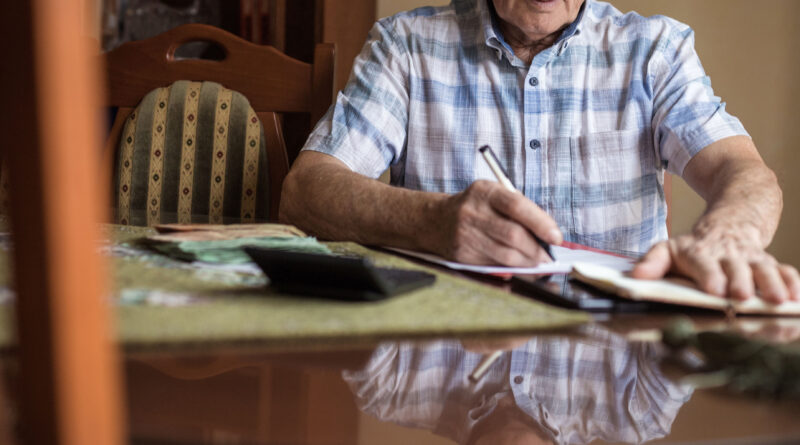 A senior man is sitting at a dining room table paying bills. He has a calculator in front of him.